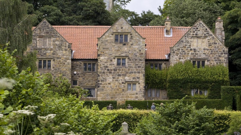 A view of a large stone manor house in the background, with long grasses and hedge in the foreground.